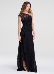 A woman modeling a floor-length black lace gown with cap sleeves and a draped neckline, photographed against a clean light gray studio background.