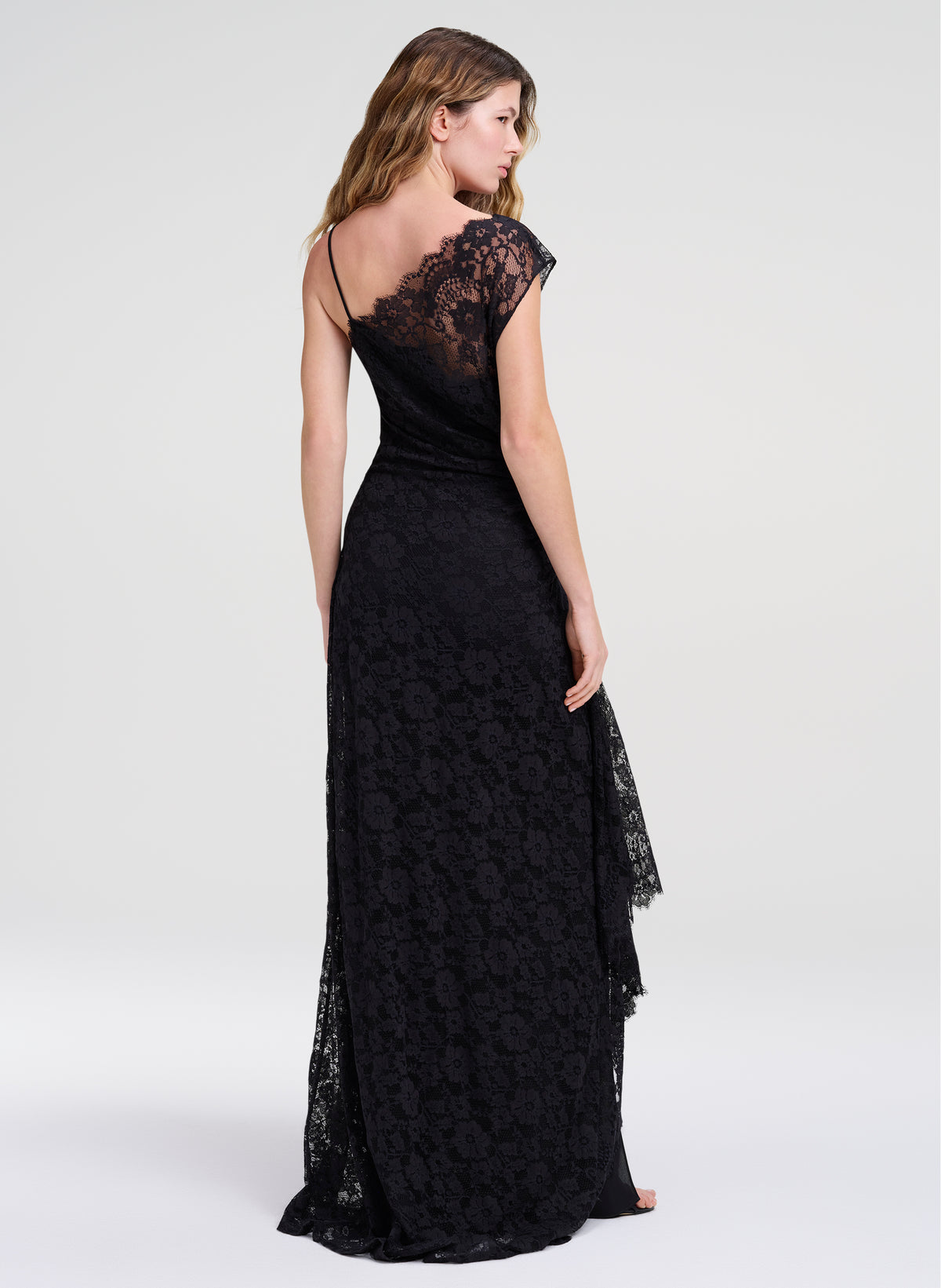 A woman modeling a floor-length black lace gown from the back, showing a one-shoulder neckline, thin straps, and a fitted skirt with a draped lace overlay on the side, against a plain light gray studio background.