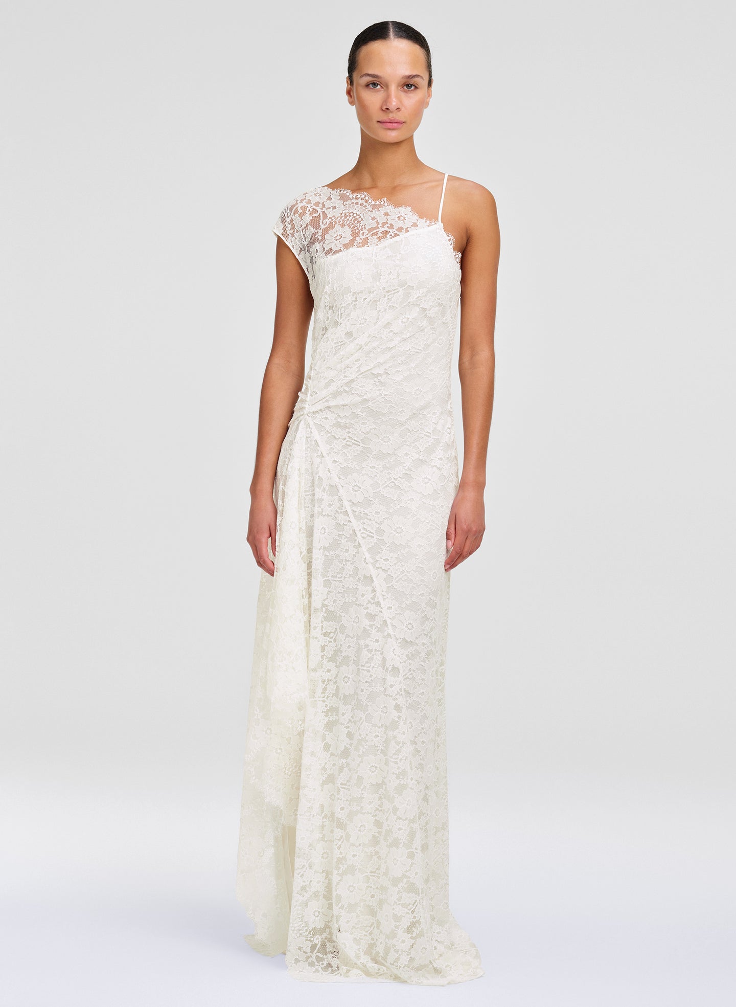 A model wearing a floor-length ivory lace gown with a one-shoulder design, cap sleeves, and thin spaghetti straps against a light gray studio background.