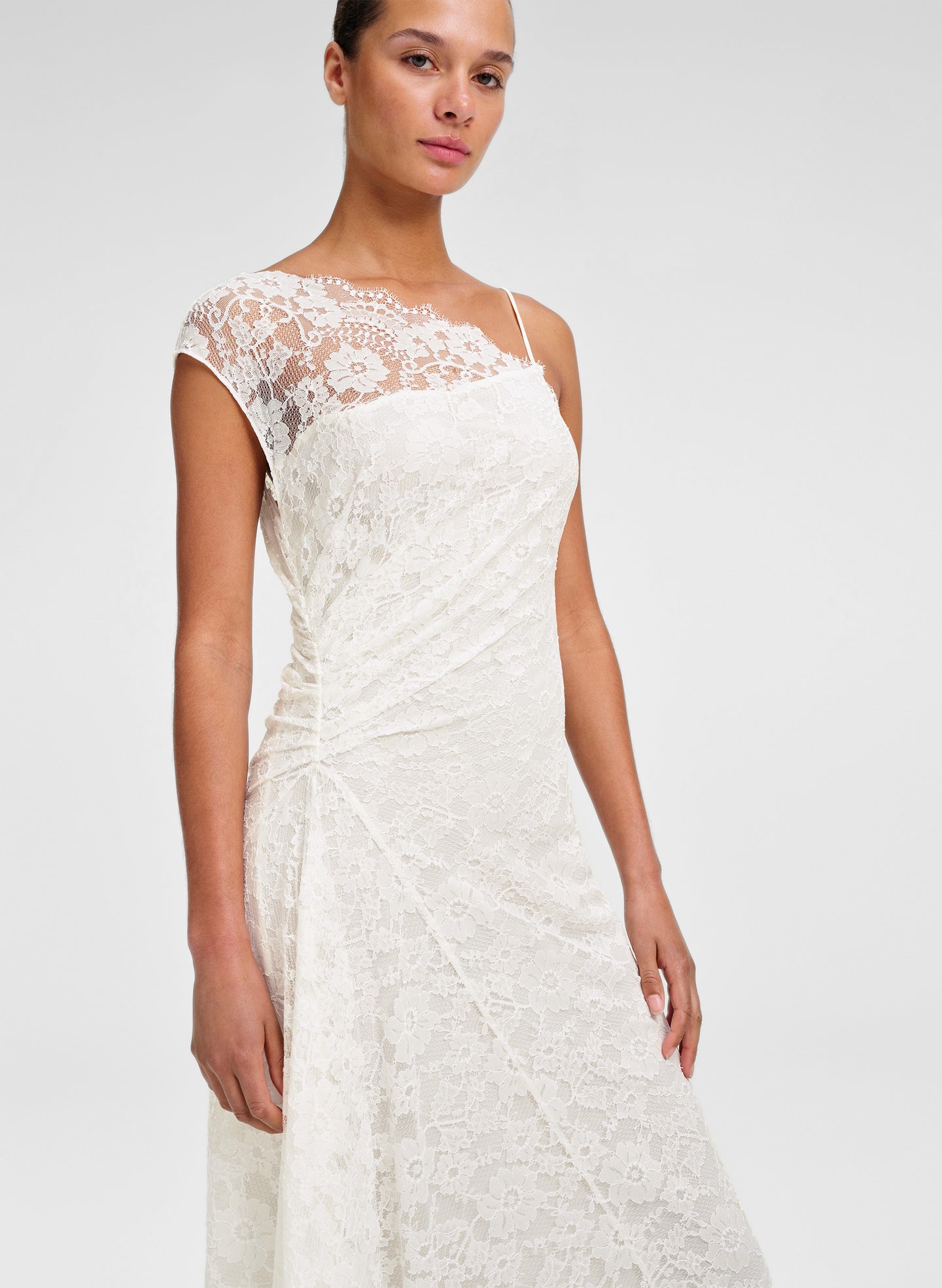 A woman modeling a white lace dress with cap sleeves, a sheer lace yoke, and thin spaghetti straps against a light gray studio background.