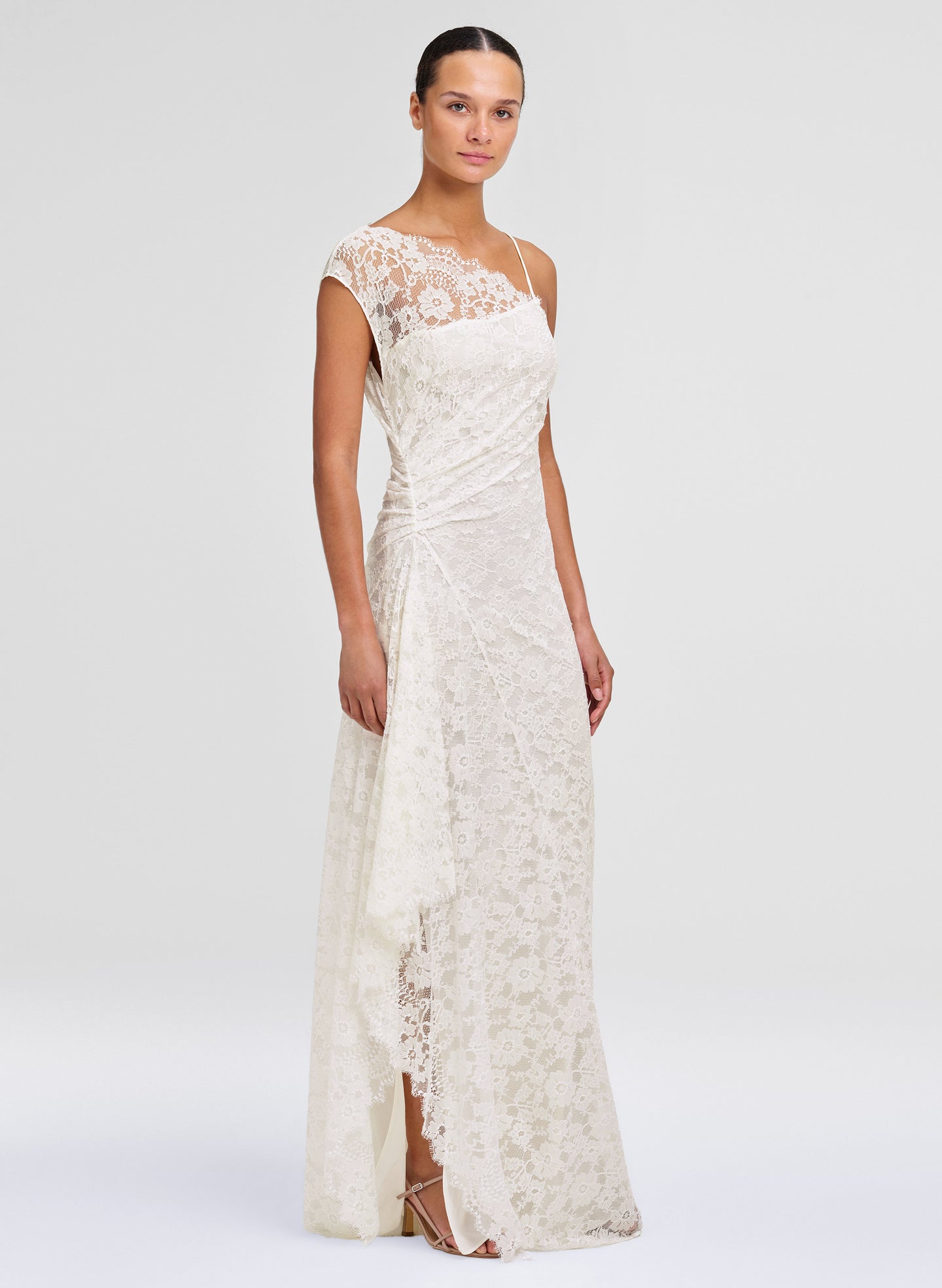 A woman modeling a floor-length ivory lace gown with cap sleeves, a draped waist, and a side slit, photographed against a clean light gray studio background.