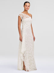A woman modeling a floor-length ivory lace gown with cap sleeves, a draped waist, and a side slit, photographed against a clean light gray studio background.
