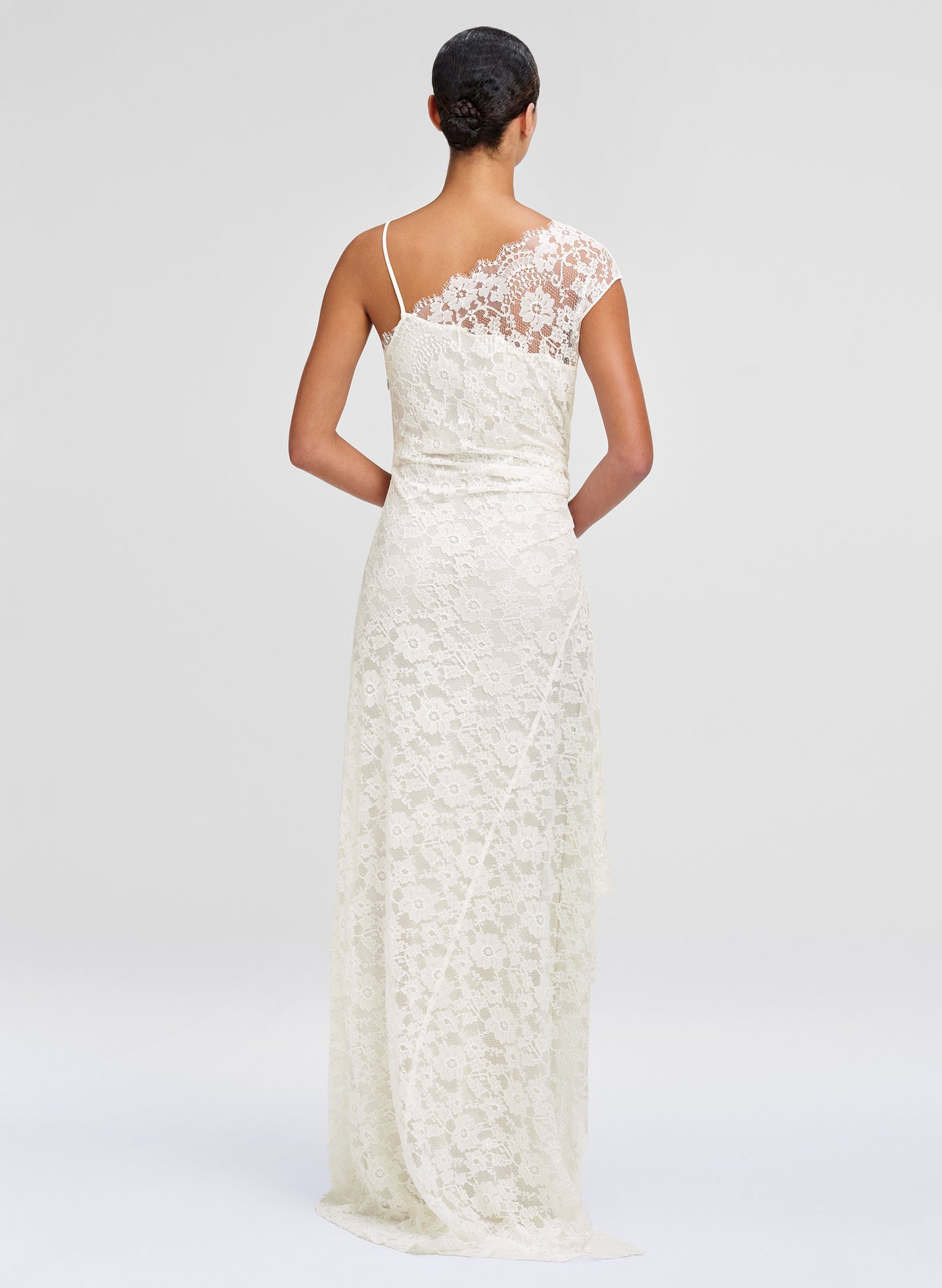 A woman shown from the back wearing a floor-length ivory lace dress with thin straps and a lace back panel, photographed against a clean light gray studio background.