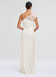 A woman shown from the back wearing a floor-length ivory lace dress with thin straps and a lace back panel, photographed against a clean light gray studio background.