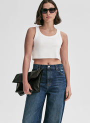 Woman wearing a white tank top and blue jeans, holding a black clutch against a plain background