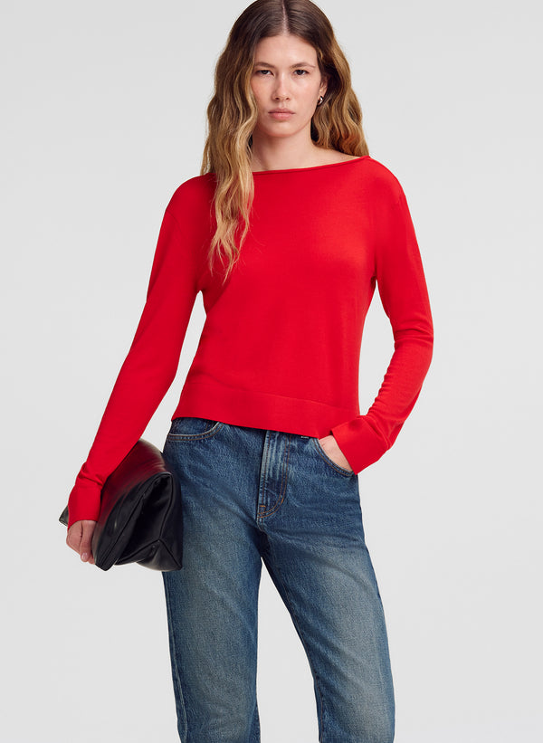 A woman modeling a bright red long-sleeve knit sweater with a wide boat neckline, paired with blue jeans and holding a black clutch bag against a light gray studio background.