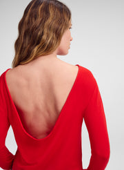 A woman shown from the back wearing a bright red long-sleeve top with a very deep open back, photographed against a light gray studio background.