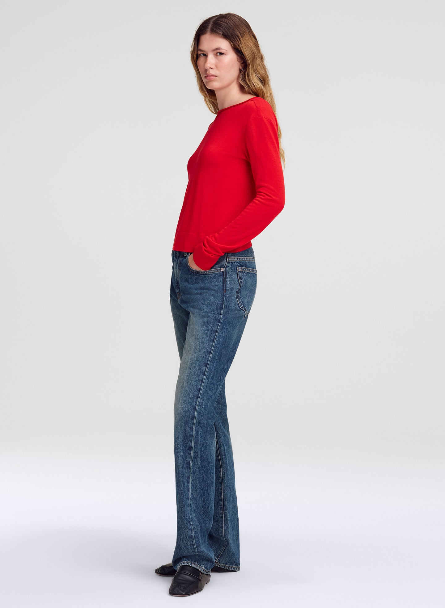 A woman modeling a bright red long-sleeve knit top and blue flare jeans against a light gray studio background.
