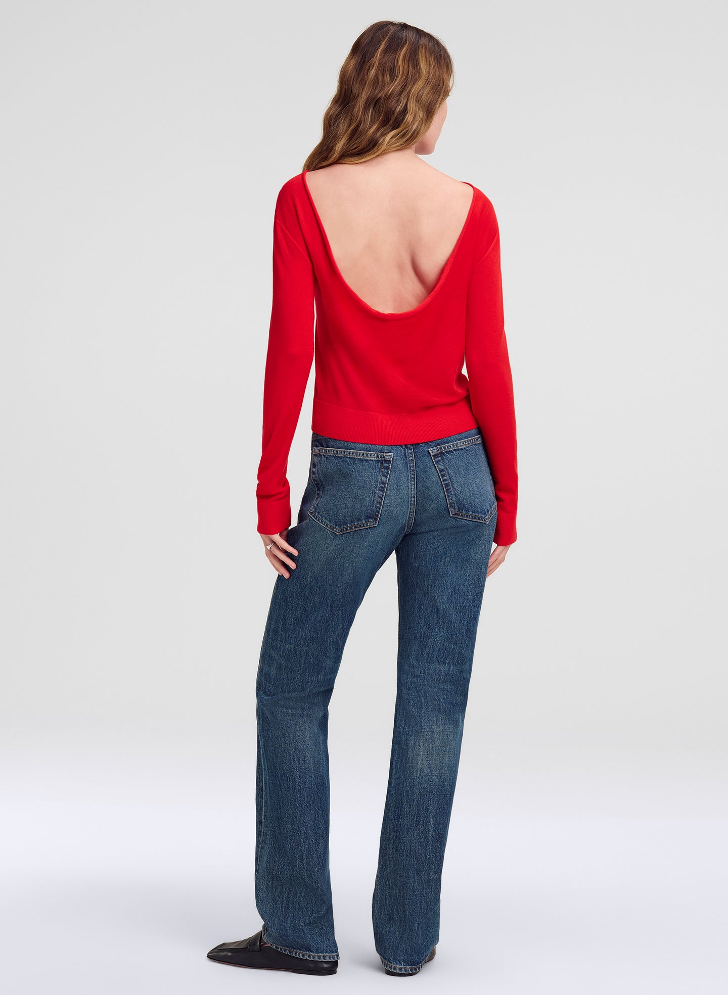 A woman shown from the back wearing a bright red long-sleeve top with a deep open back and blue jeans, standing against a light gray studio background.