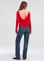 A woman shown from the back wearing a bright red long-sleeve top with a deep open back and blue jeans, standing against a light gray studio background.