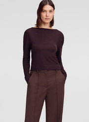 A woman modeling a dark brown long-sleeve knit top with a wide boat neckline, paired with brown trousers, against a light gray studio background.