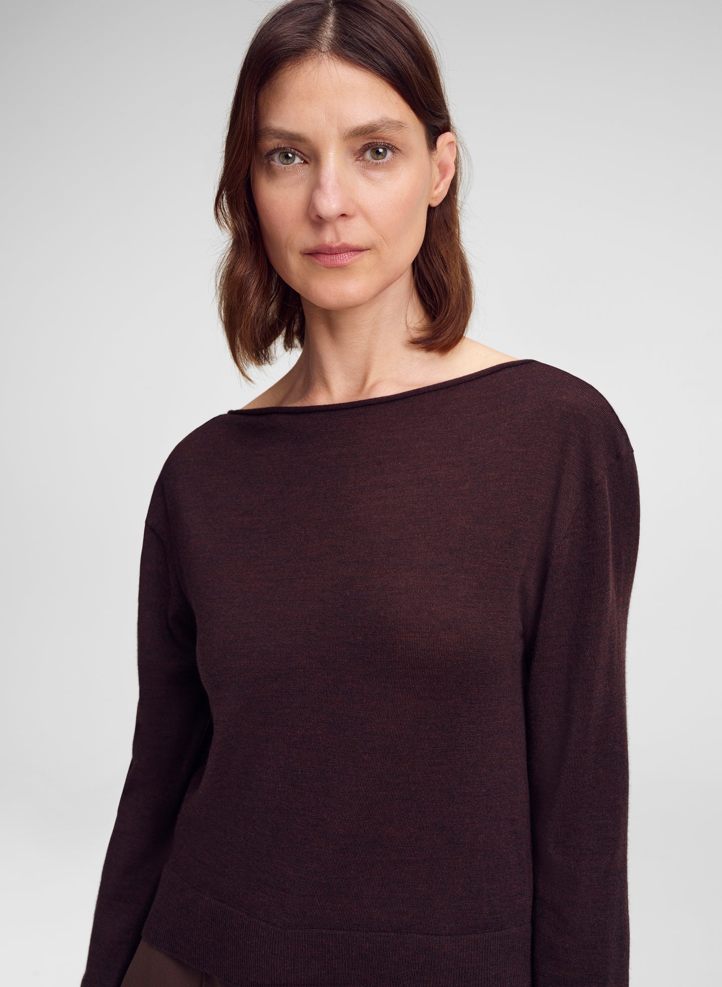 A woman wearing a dark brown wide-neck long-sleeve knit top, photographed against a neutral light gray studio background.