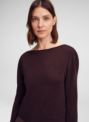 A woman wearing a dark brown wide-neck long-sleeve knit top, photographed against a neutral light gray studio background.