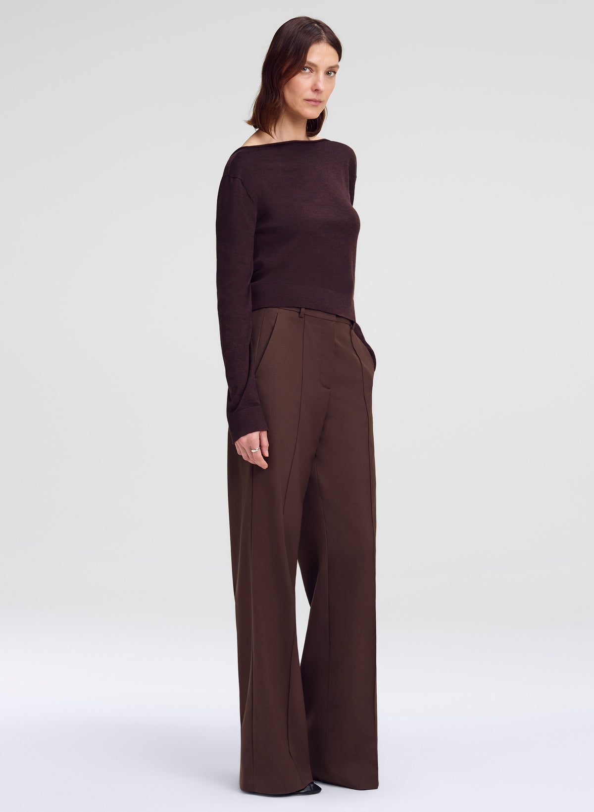 A woman modeling a dark brown off-the-shoulder long-sleeve top paired with matching dark brown wide-leg trousers, photographed against a seamless light gray studio background.