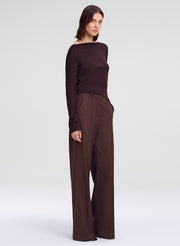 A woman modeling a dark brown off-the-shoulder long-sleeve top paired with matching dark brown wide-leg trousers, photographed against a seamless light gray studio background.