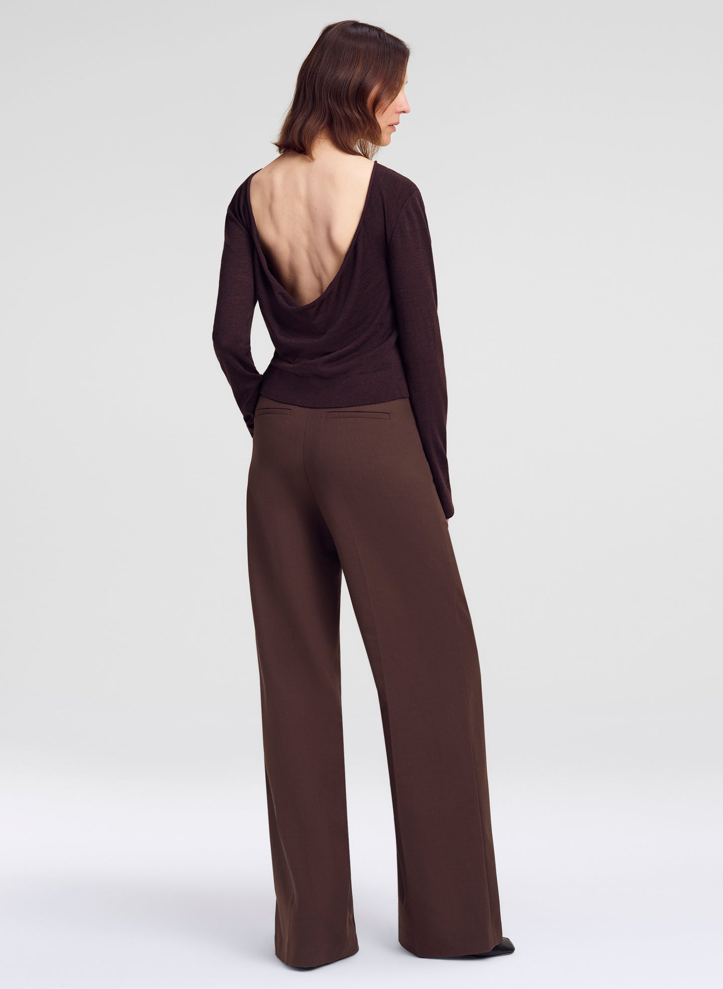 A model shown from the back wearing a dark brown long-sleeve top with a dramatic open back and matching wide-leg trousers, set against a plain light gray studio background.