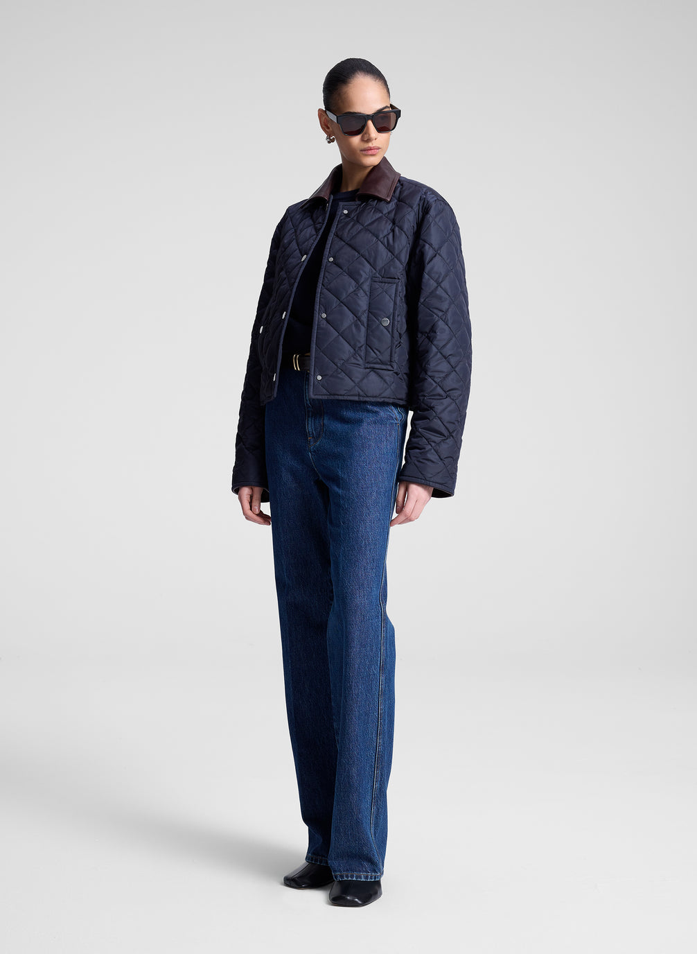 Person wearing a navy quilted jacket and sunglasses on a light gray background