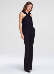 A woman modeling a black sleeveless lace halter top with matching black wide-leg trousers, photographed against a clean light gray studio background.