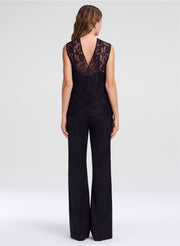 A woman viewed from the back wearing a sleeveless black lace top with a deep V back and wide-leg pants, photographed against a light gray studio background.