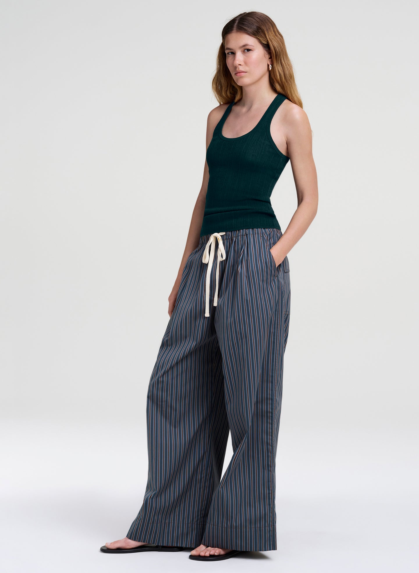 A woman modeling a dark green ribbed tank top and wide-leg pinstriped pants with a cream drawstring, standing against a light gray studio background.