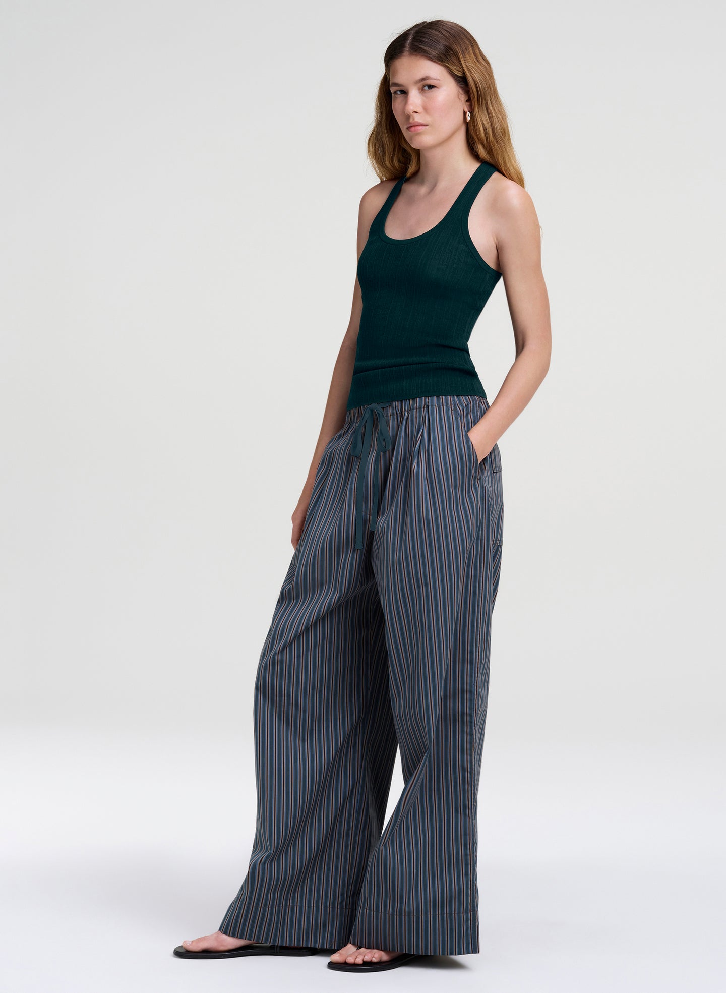 A woman modeling a dark green ribbed tank top and wide-leg pinstriped trousers in a studio setting with a light gray background.