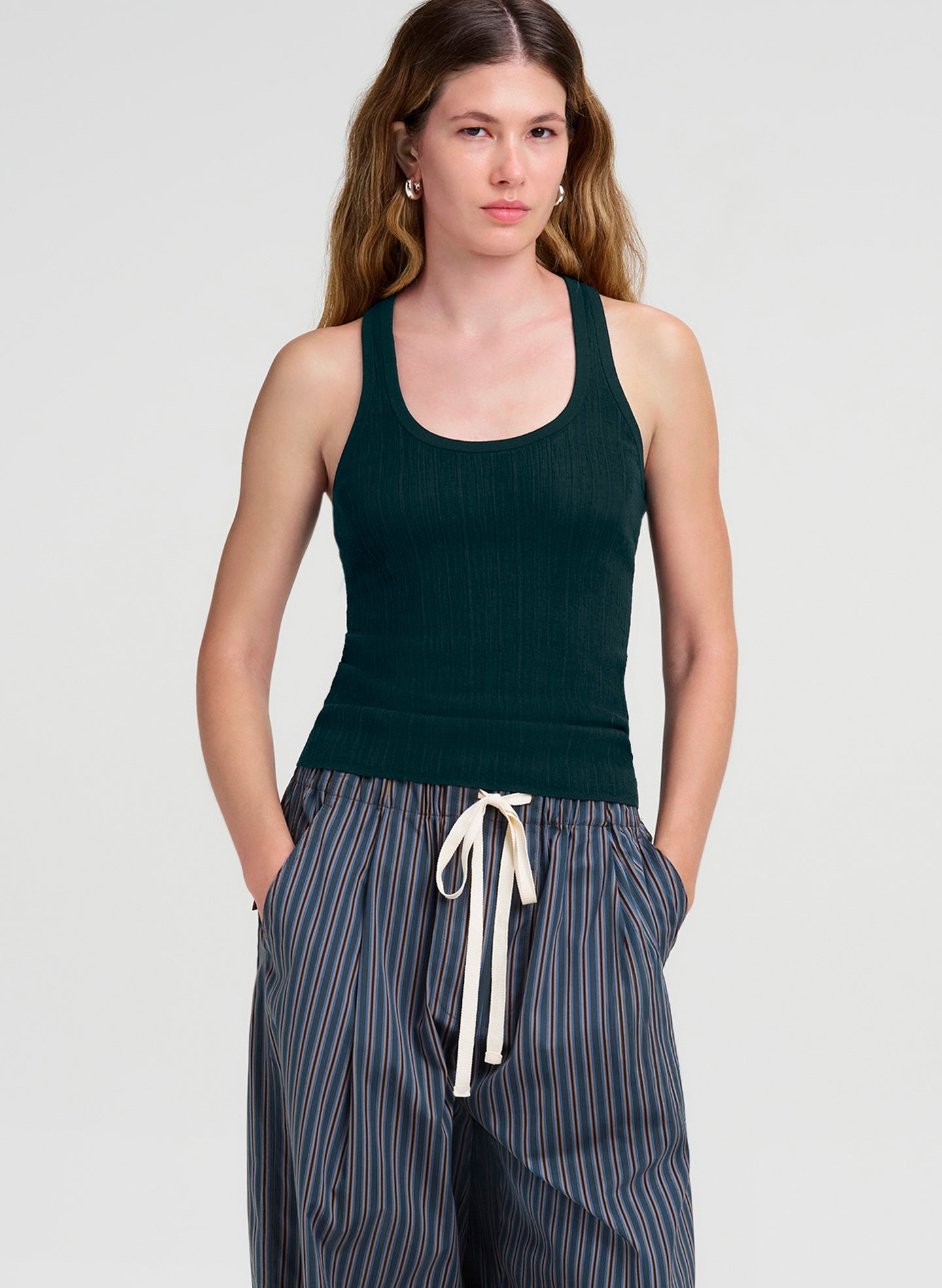 A woman modeling a dark green ribbed tank top and blue-and-white striped drawstring pants against a light gray studio background.