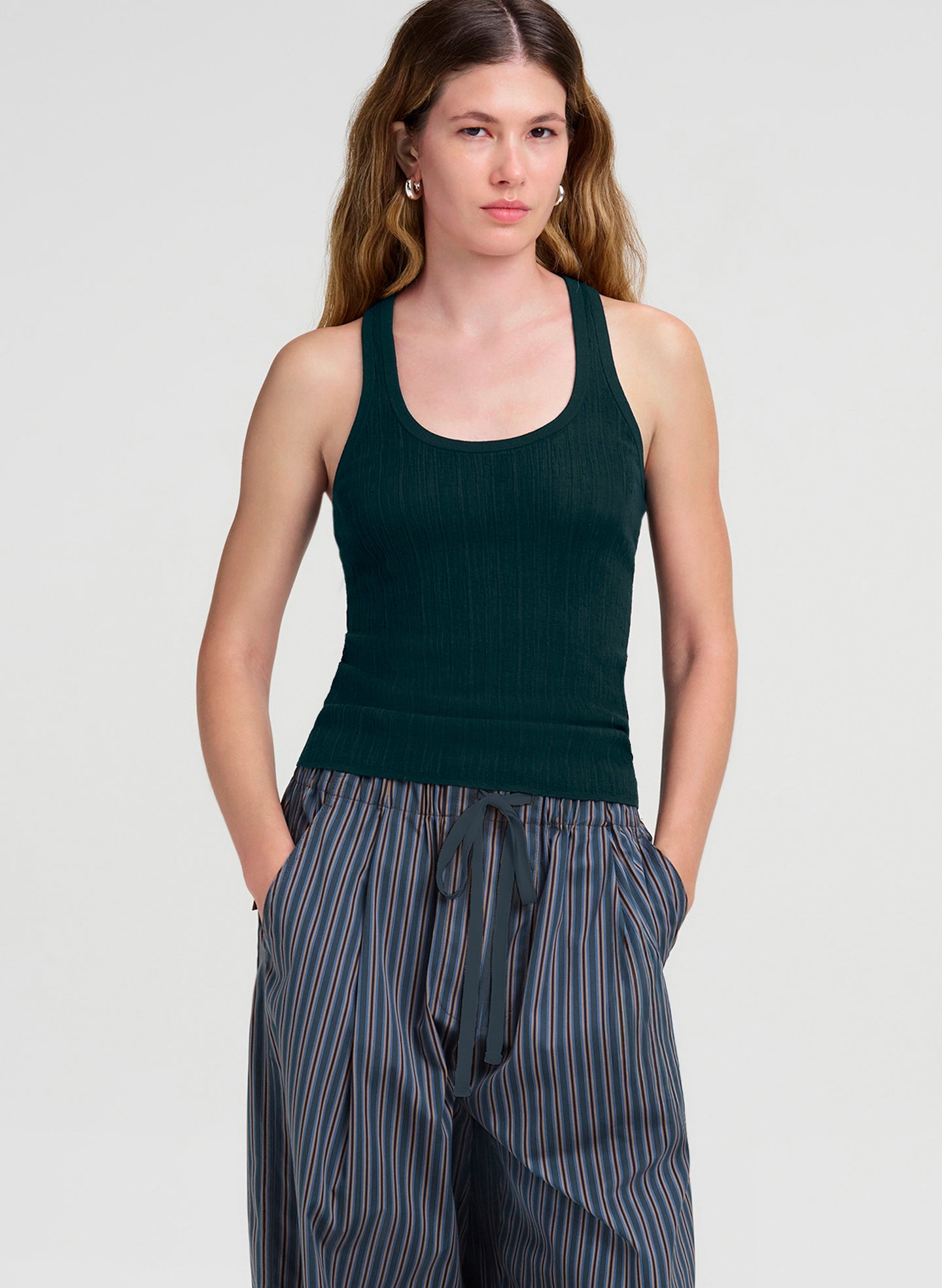 A woman modeling a dark green ribbed tank top paired with blue and gray vertically striped drawstring pants against a light gray studio background.