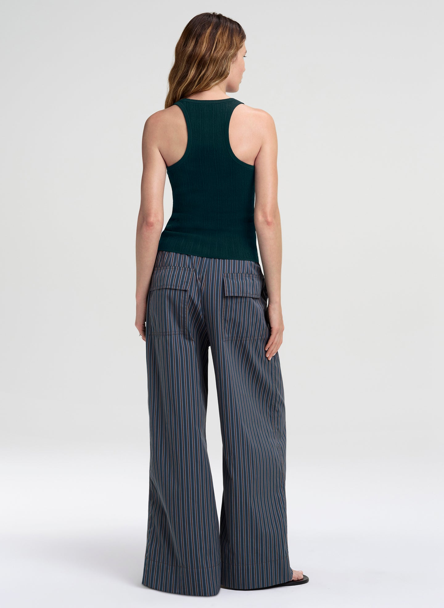 A woman shown from the back wearing a dark green ribbed tank top and slate blue wide-leg pinstripe pants with thin orange stripes, set against a plain light gray studio background.