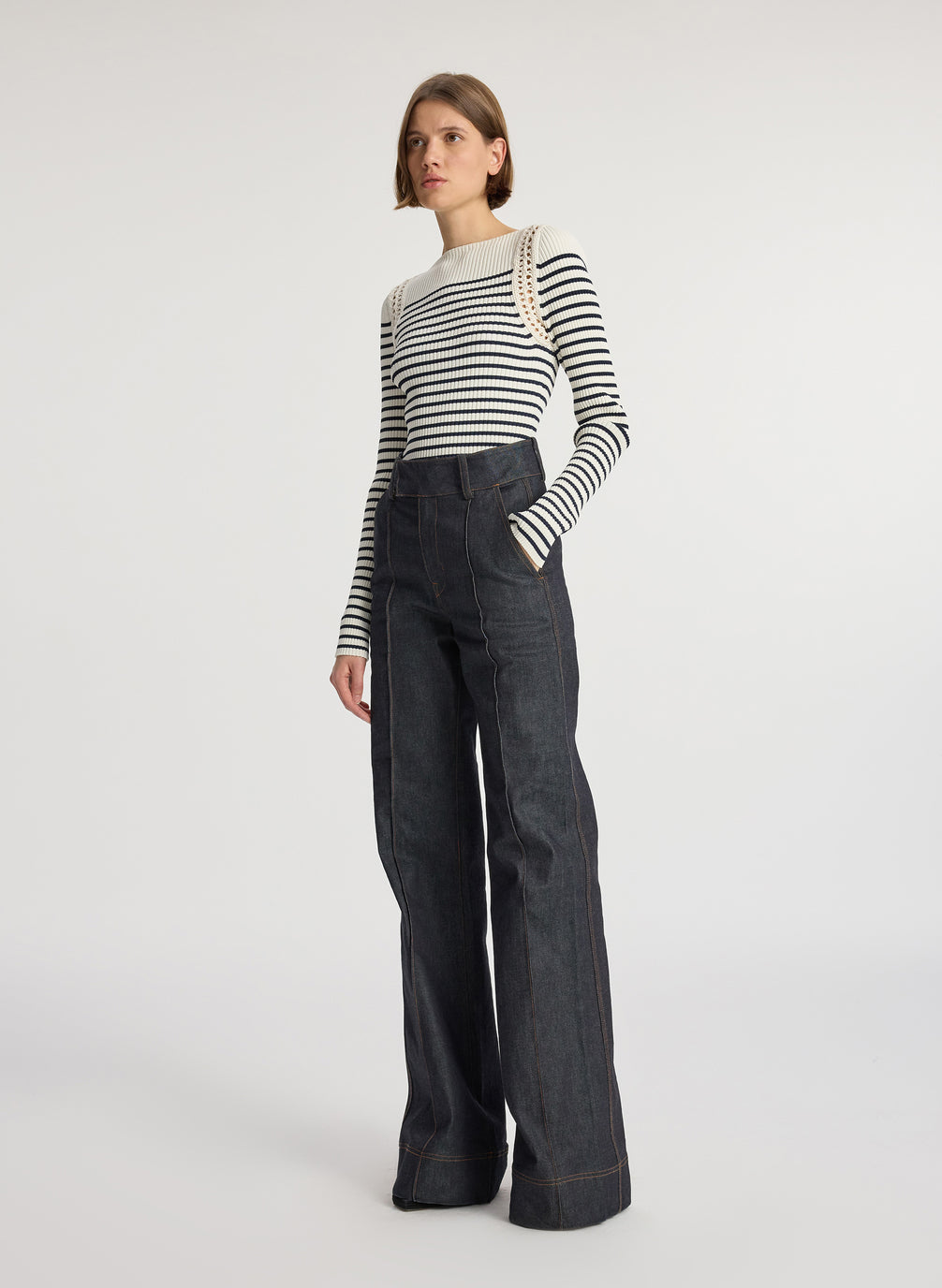 side view of woman wearing white and navy striped top with long bell sleeves and dark wash raw denim jeans