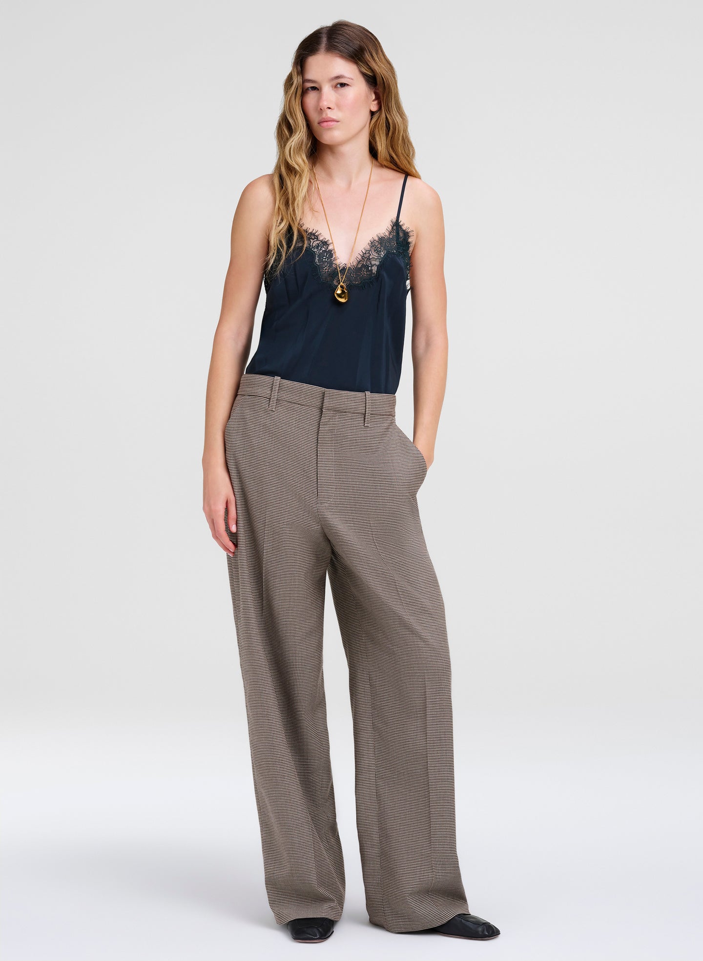 A woman modeling a dark green camisole with lace trim and wide-leg taupe trousers against a light gray studio background.
