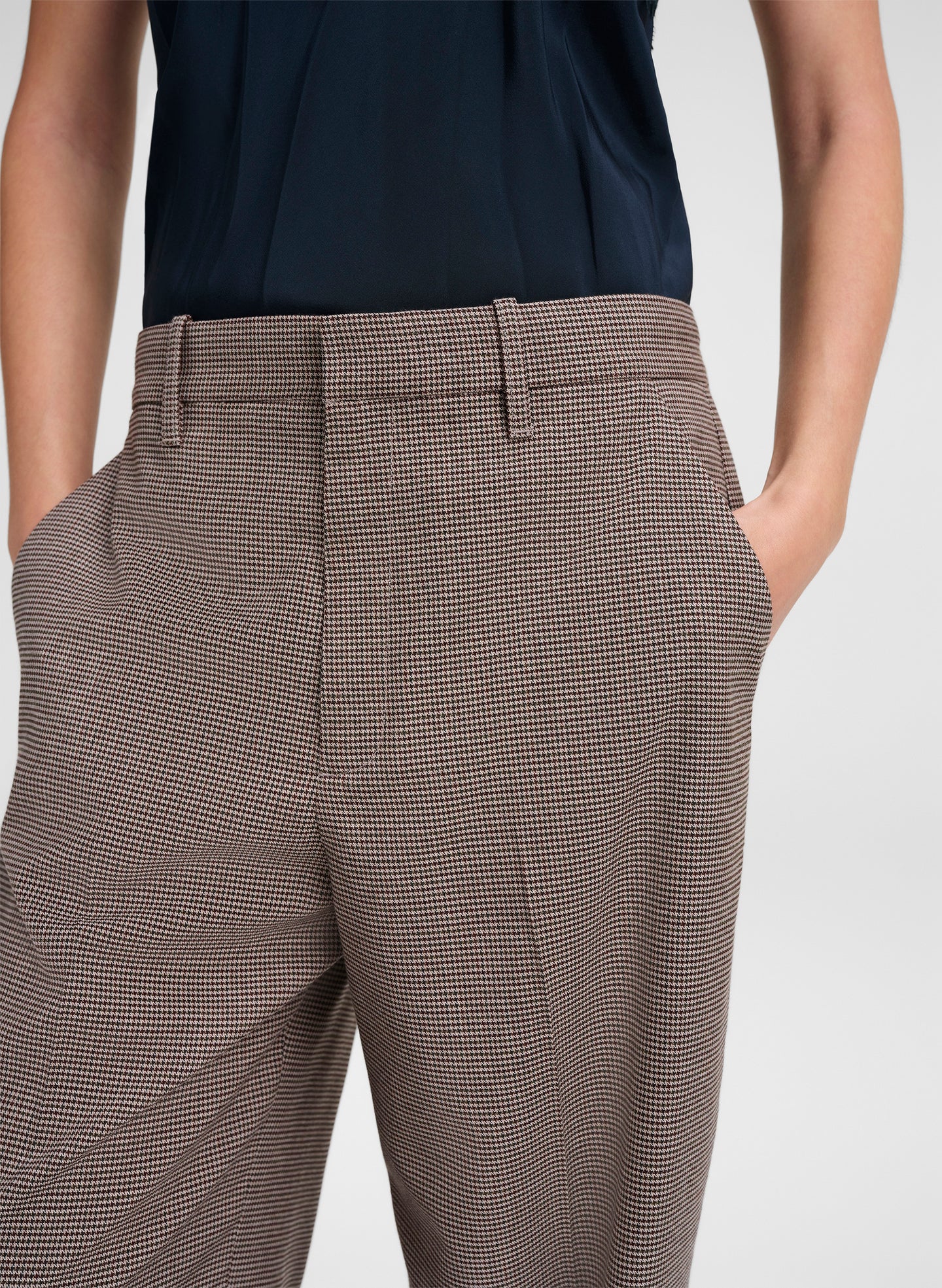 Close-up of a person wearing trousers with a fine brown and beige houndstooth pattern, paired with a dark navy top against a light gray studio background.