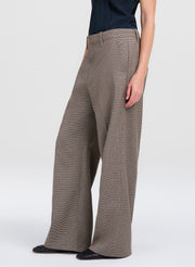 A model wearing a pair of wide-leg trousers in a textured taupe-brown fabric, paired with a dark top and black shoes, shown from the waist down on a light gray studio background.