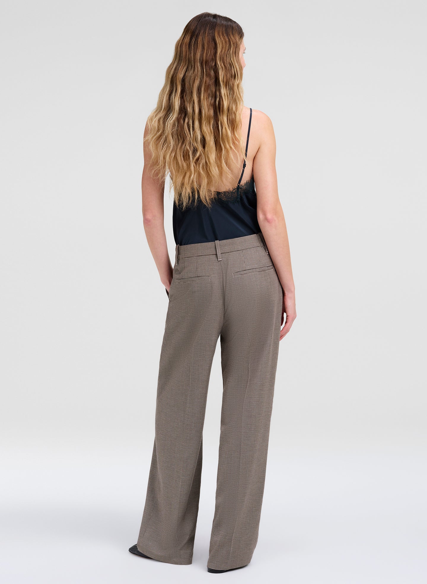 A woman viewed from behind wearing high-waisted, wide-leg taupe trousers and a dark green spaghetti-strap camisole top, set against a plain light gray studio background.