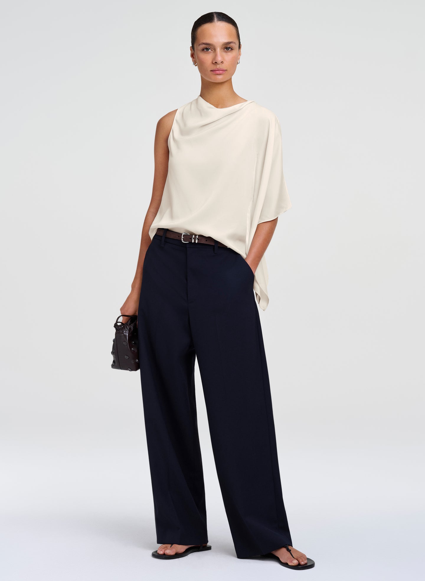 A model stands against a light gray studio background wearing a cream draped sleeveless top, dark navy wide-leg trousers, a thin black belt, black sandals, and holding a small black handbag.
