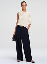 A model stands against a light gray studio background wearing a cream draped sleeveless top, dark navy wide-leg trousers, a thin black belt, black sandals, and holding a small black handbag.
