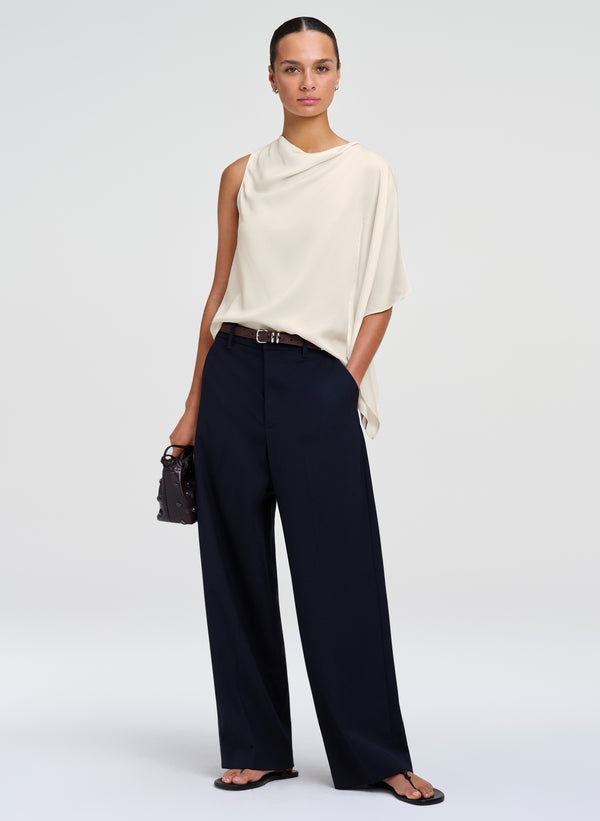 A model stands against a light gray studio background wearing a cream draped sleeveless top, dark navy wide-leg trousers, a thin black belt, black sandals, and holding a small black handbag.
