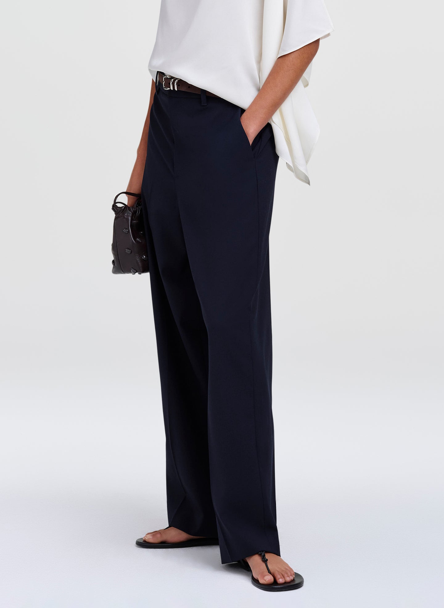 A model wearing navy blue wide-leg trousers, styled with a white top, black sandals, and a small dark brown handbag against a light gray studio background.