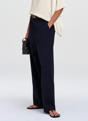 A model wearing navy blue wide-leg trousers, styled with a cream-colored blouse, a black belt, and black sandals, holding a small black handbag against a light gray studio background.