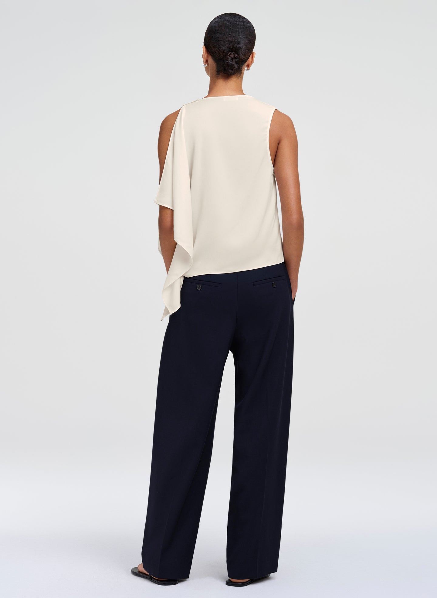 A model shown from the back wearing a white sleeveless draped top and dark navy wide-leg trousers, photographed against a seamless light gray studio background.