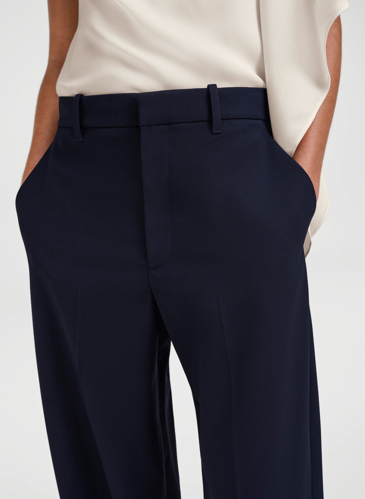 Close-up of a person wearing navy blue tailored trousers with a cream blouse, shown from the chest to the upper thighs against a light gray studio background.