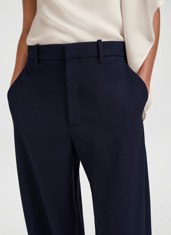 Close-up of a person wearing navy blue tailored trousers with a cream blouse, shown from the chest to the upper thighs against a light gray studio background.
