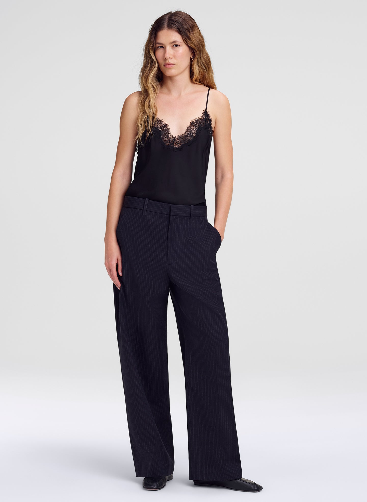 A woman modeling a black camisole top with lace trim and black wide-leg trousers against a light gray studio background.