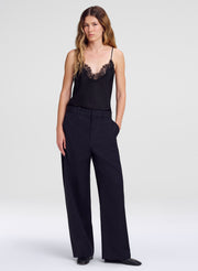 A woman modeling a black camisole top with lace trim and black wide-leg trousers against a light gray studio background.