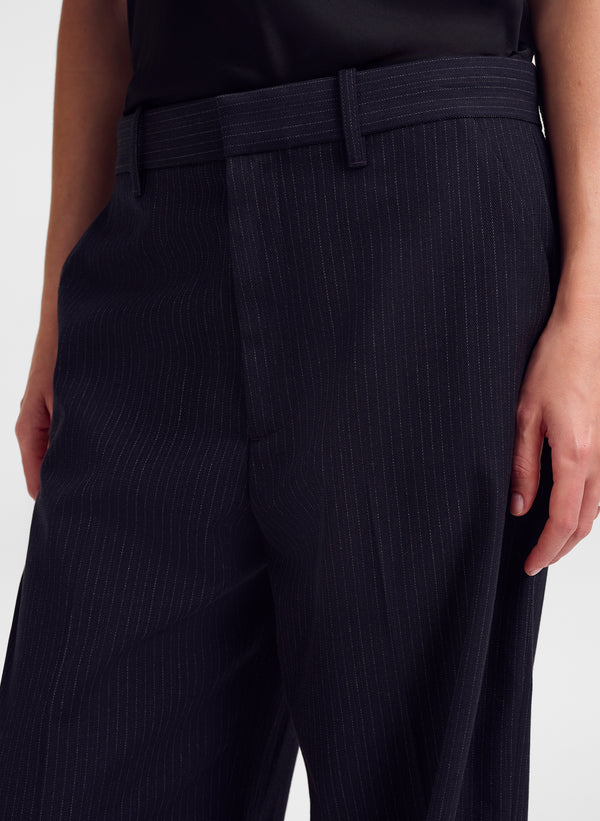 Close-up of a person wearing dark navy pinstriped trousers with a matching belt, paired with a black top against a light gray studio background.