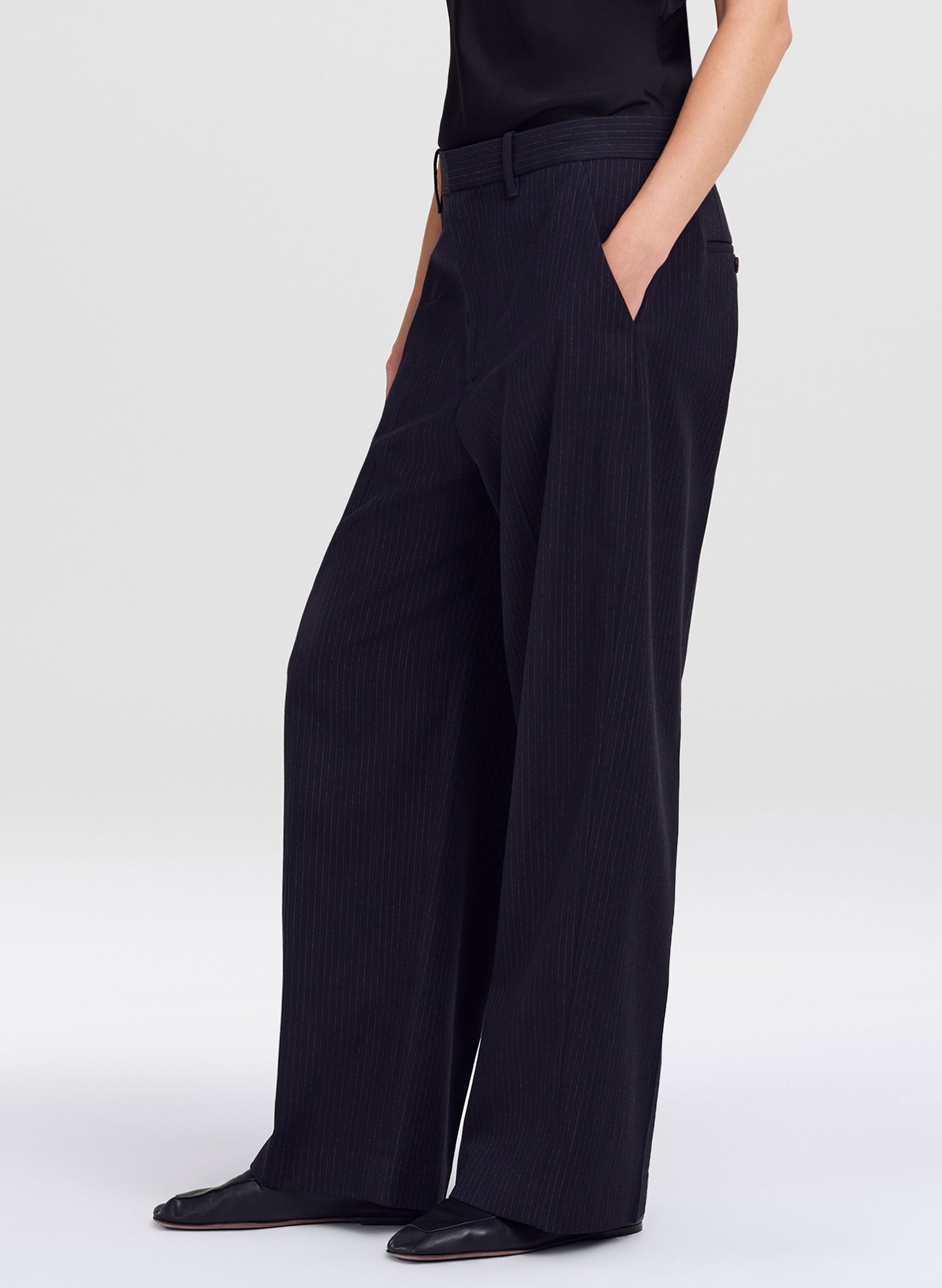 A model wearing dark navy blue wide-leg trousers with subtle vertical pinstripes, paired with a black top and black shoes, shown from the waist down against a light gray studio background.