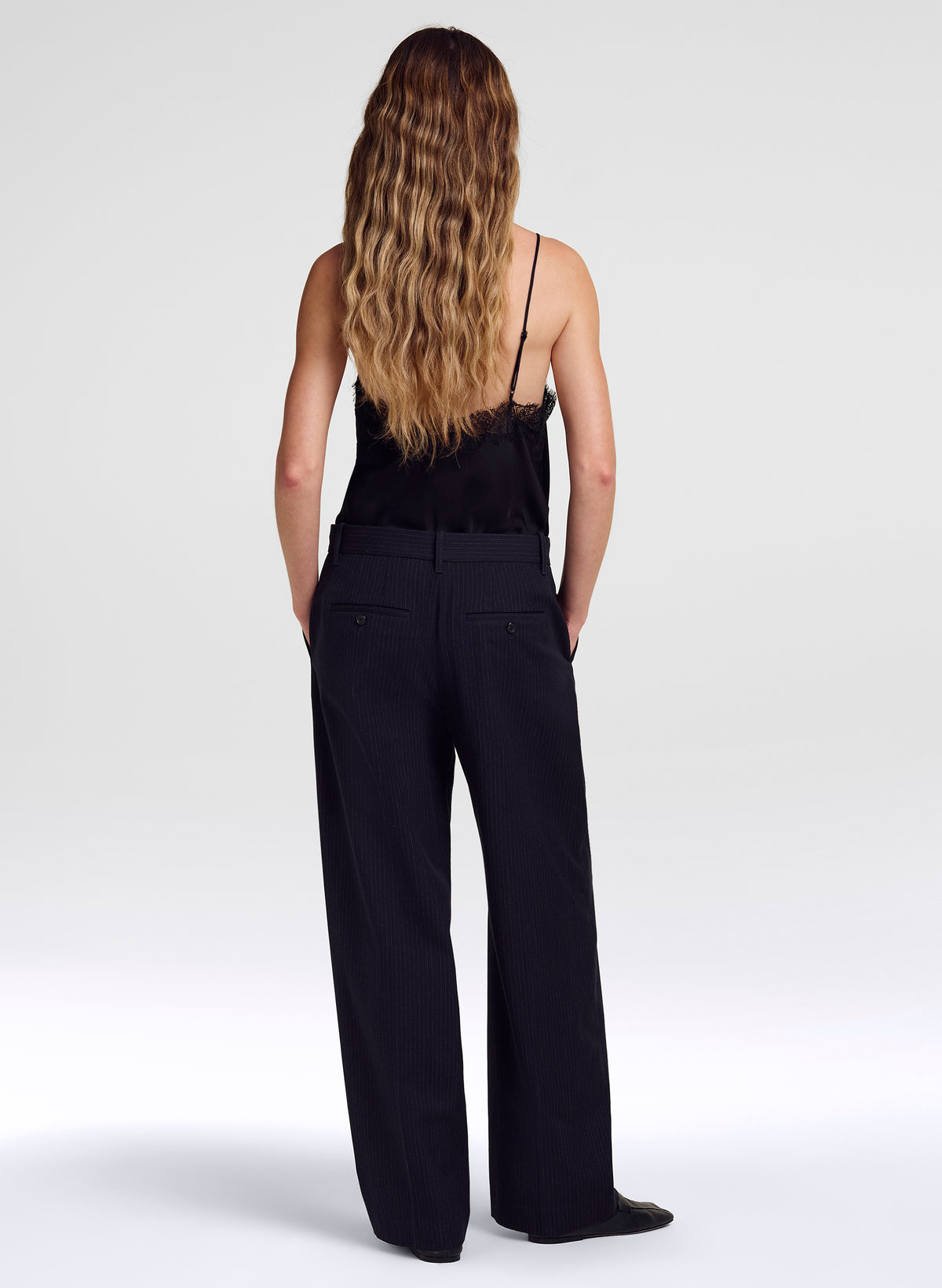 A woman with long wavy light brown hair is shown from behind wearing a black camisole and black wide-leg trousers against a light gray studio background.