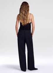 A woman with long wavy light brown hair is shown from behind wearing a black camisole and black wide-leg trousers against a light gray studio background.