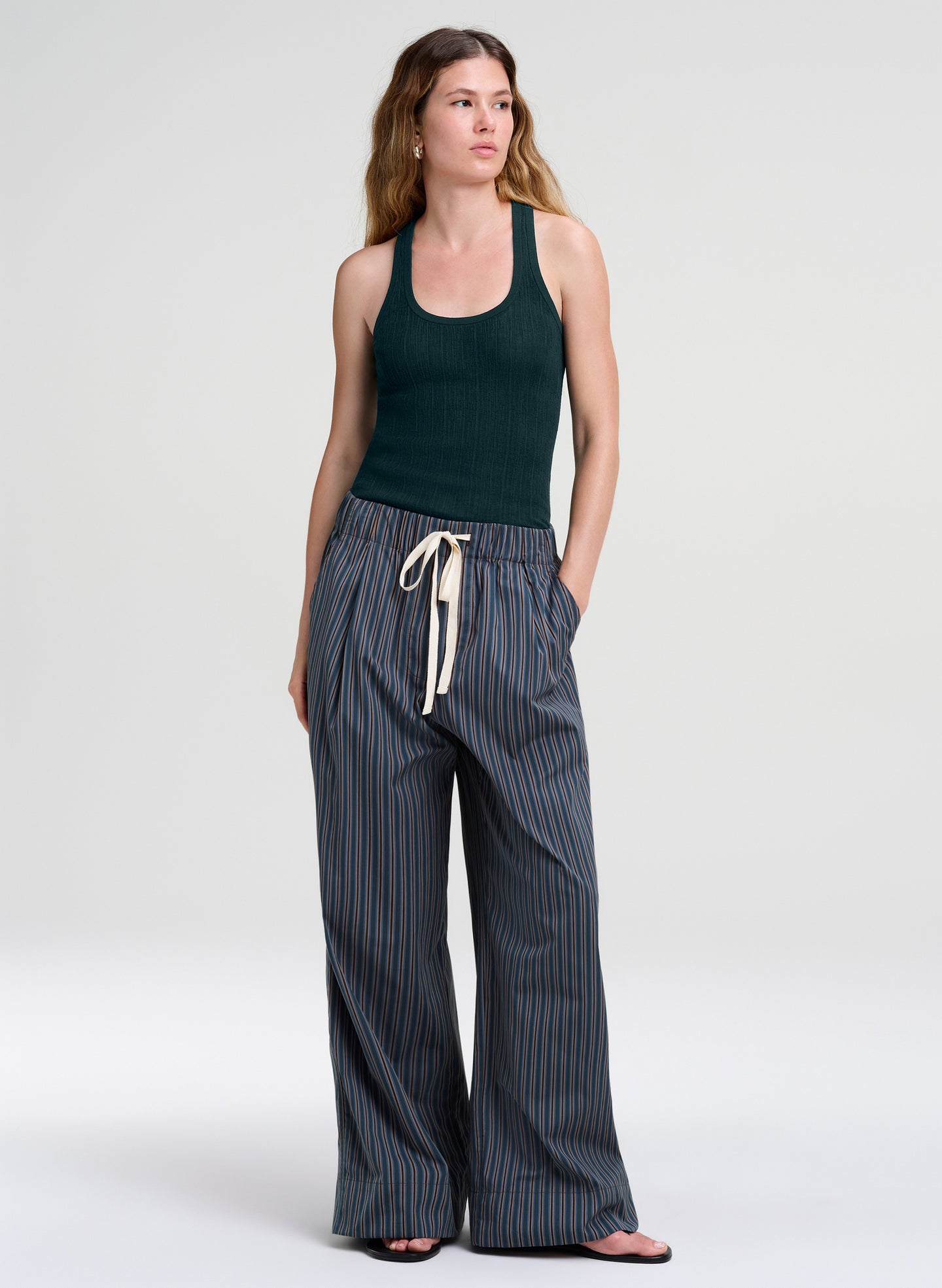 A woman modeling a dark green ribbed tank top and wide-leg striped pants with a cream drawstring, standing against a light gray studio background.