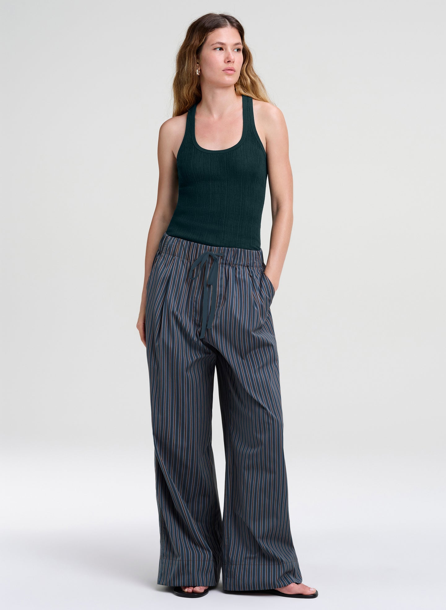 A woman modeling a dark green ribbed tank top and wide-leg pinstriped trousers in a studio setting with a light gray background.