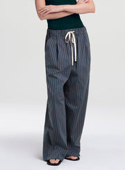 A model wearing wide-leg trousers with vertical navy and gray stripes, featuring an elastic waistband and a cream-colored drawstring, paired with a dark green top and black sandals.